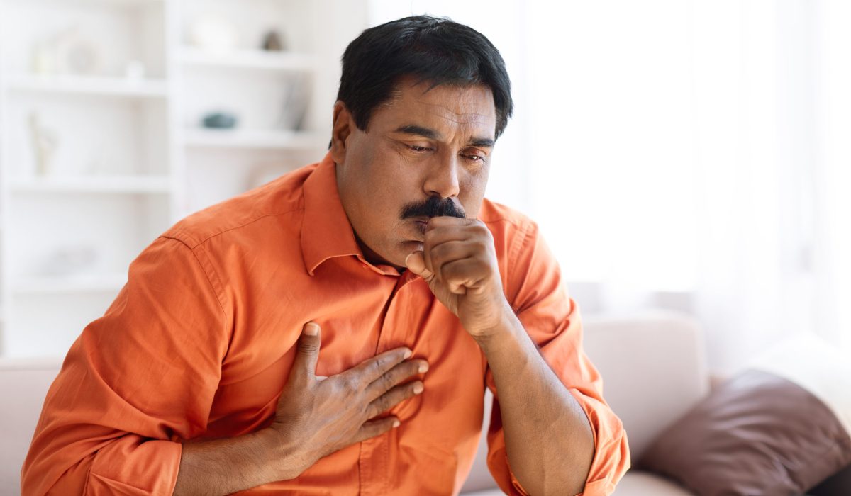 Chronic cough, reasons and treatment. Unhealthy sick indian middle aged man wearing orange shirt sitting on couch and coughing, touching chest, home interior, copy space